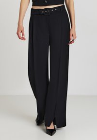 Frau in hoch taillierten schwarzen, weit geschnittenen Hosen mit einem Gürtel und seitlichen Schlitzen, kombiniert mit schwarzen spitzen Schuhen auf einem neutralen Hintergrund.