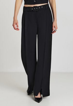 Woman wearing high-waisted black wide-leg trousers with a belt and side slits, paired with black pointed-toe shoes on a neutral background.