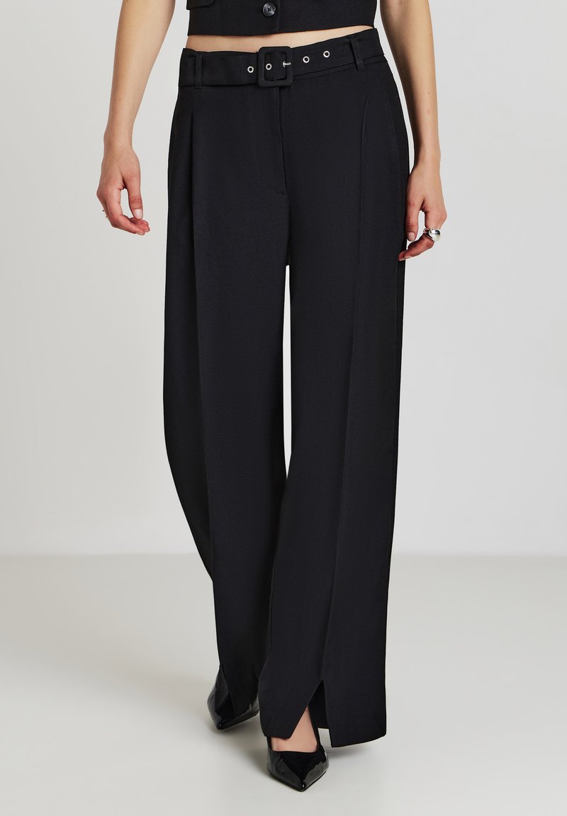 Frau in hoch taillierten schwarzen, weit geschnittenen Hosen mit einem Gürtel und seitlichen Schlitzen, kombiniert mit schwarzen spitzen Schuhen auf einem neutralen Hintergrund.