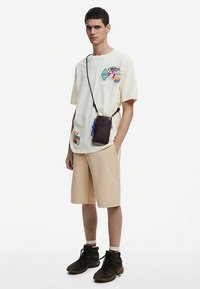 Svijetložuta košulja kratkih rukava s grafičkim zakrpama, bež kratke hlače i smeđa torba preko ramena. Crne čizme sa crvenim i bijelim prugastim čarapama.