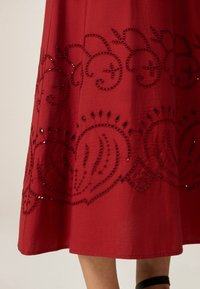 Jupe rouge avec un motif découpé complexe et des détails en perles sur l'ourlet. Tissu lisse avec une légère brillance, s'évasant doucement en bas.