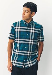 Junger Mann mit einem kurzärmeligen Hemd mit kariertem Muster in Grün, Weiß und Marineblau, der nach links blickt und eine Hand in der Tasche hat.