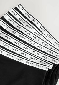Črne legice z belim elastičnim pasom, ki vsebujejo ponovljen napis "NEXT." Material se zdi mehak in gladek ter natančno prilega.