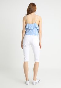 Vaaleansininen röyhelötoppi kukkakuviolla, yhdistetty valkoisiin lyhyisiin pantsareihin. Mallilla on päällään valkoiset lenkkitossut, kuvattu takaa.