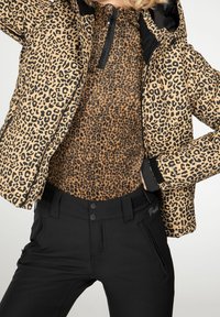 Personne portant une veste imprimé léopard et un top zippé assorti imprimé léopard, associé à un pantalon noir taille haute avec des poches zippées.