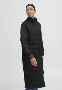 Abrigo largo acolchado negro con cuello alto, que cuenta con un cierre de botones y bolsillos laterales. Fabricado con una tela suave y texturada.