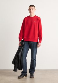 Rød bomulds sweatshirts med ribbede manchetter og kant, iført mørkeblå jeans. Modellen holder en sort læderjakke og har sorte sko på.