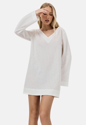 Blonde vrouw draagt een losse, witte, lange mouwen V-hals jurk en raakt met haar rechterhand haar voorhoofd aan tegen een effen achtergrond.