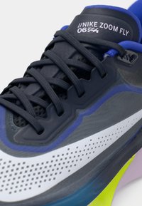 Las zapatillas Nike Zoom Fly cuentan con una parte superior en negro y gris con acentos en azul, textura de malla, cierre de cordones y un distintivo diseño de swoosh blanco.