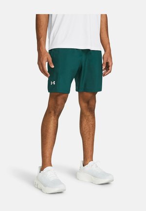 Man draagt donkergroene Under Armour korte broek, witte sportschoenen en een wit shirt, staand tegen een witte achtergrond.
