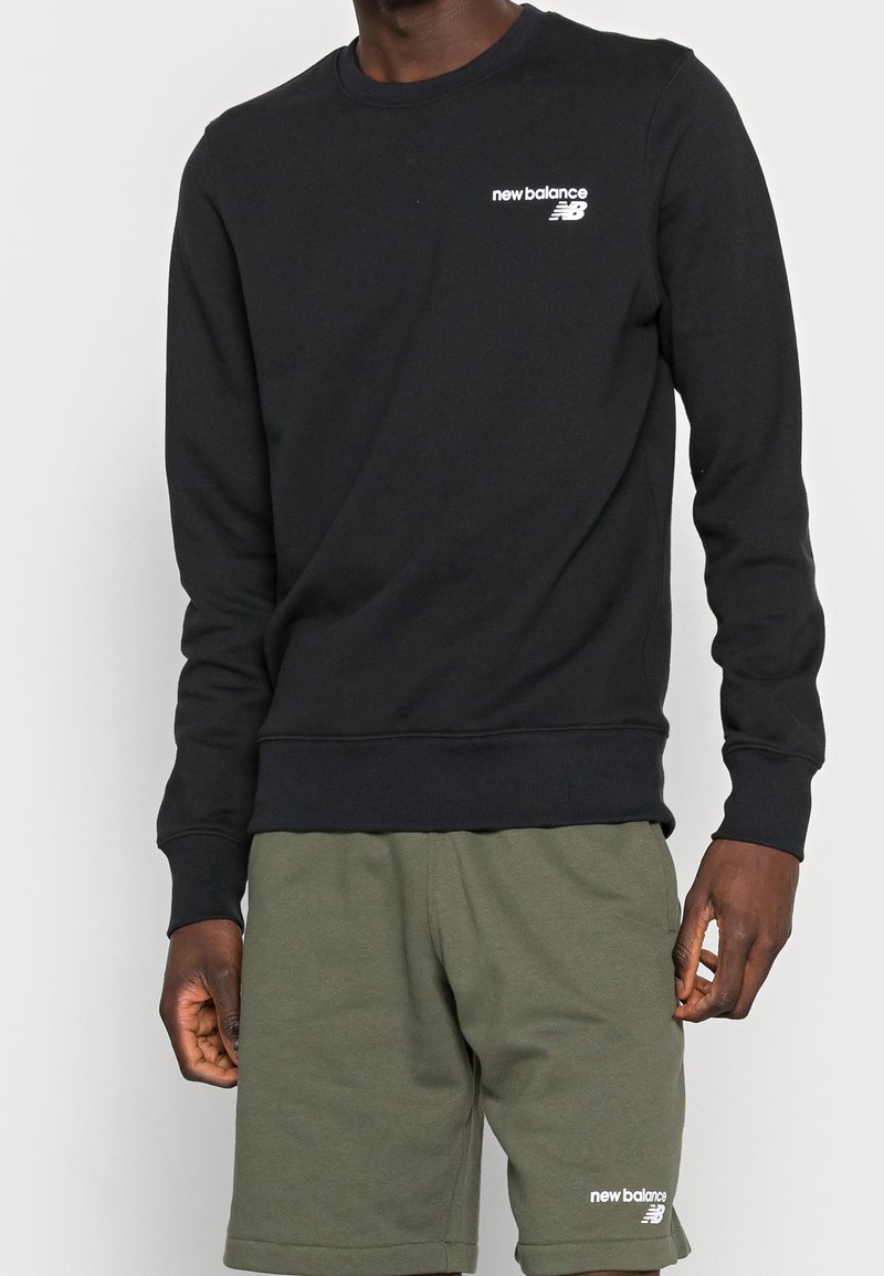Homme portant un sweat-shirt New Balance noir et un short New Balance vert olive sur un fond uni.
