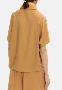 Kobieta w luźnej, krótkiej bluzce w kolorze kamela z kołnierzykiem oraz pasujących szerokich spodniach, widoczna z tyłu.
