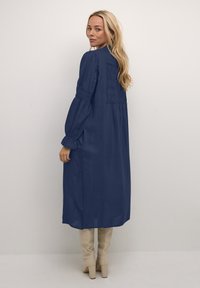 Abito lungo blu navy con schiena arricciata, maniche a palloncino e silhouette fluida. Abbinato a stivali beige fino al ginocchio.
