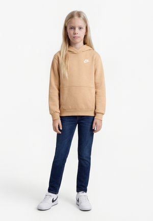 Young girl with long blonde hair wearing a beige Nike hoodie, dark blue jeans, and white Nike sneakers, standing against a white background.