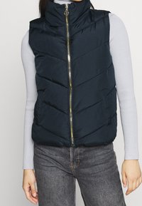 Granatowa kamizelka puchowa z wysokim kołnierzem, zapinana na zamek, z dodatkami w kolorze złotym. Posiada przeszycia w kształcie szewronu oraz boczne kieszenie. Miękka, pikowana faktura.