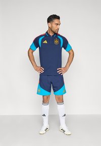 Tmavomodrý športový dres s červenými a tyrkysovými akcentmi, so zapínaním na zips a emblémom Španielska. Zodpovedajúce šortky v tmavomodrej a tyrkysovej farbe.