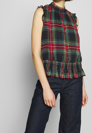 Vrouw draagt een mouwloze rood, groen, zwart en geel geruite top met gerimpelde zoom en donkerblauwe spijkerbroek tegen een effen achtergrond.