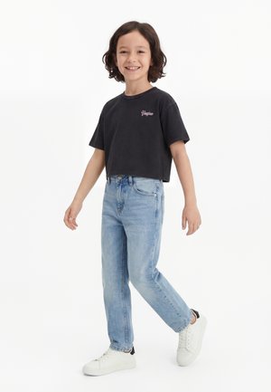 Smilende barn med middels langt mørkt hår som har på seg en svart cropped T-skjorte, lyseblå jeans og hvite joggesko, stående mot hvit bakgrunn.