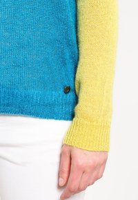 Stickad tröja med blå kropp och gula ärmar, med en strukturerad yta och en liten metalldetalj med logotyp nära fållen.