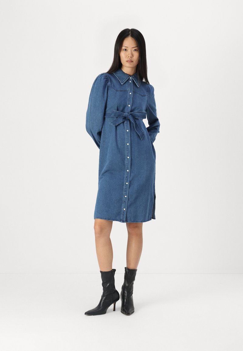 Denim jurk met een knoopsluiting aan de voorkant, lange mouwen en een strik in de taille. Heeft een kraag, zijsplitten en een lichtblauwe wassing. Zwarte enkellaarsjes.