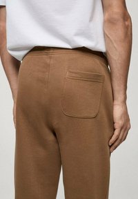 Pantaloni de trening maro cu o textură moale și netedă și o pungă la spate. Combinați cu un tricou alb simplu, evidențiind un aspect casual.