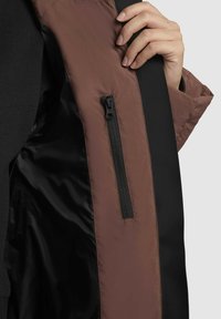 Barna és fekete dzseki belső rész, sima textúrával. Bal oldalon cipzáras zseb található a biztonságos tárolás érdekében.