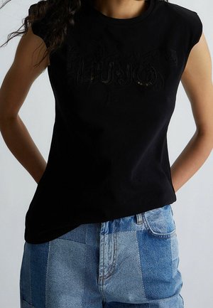 Kobieta ubrana w czarną koszulkę bez rękawów z subtelnym napisem i jeansy z niebieskiego denimu w stylu patchwork, ręce spoczywają za jej plecami.