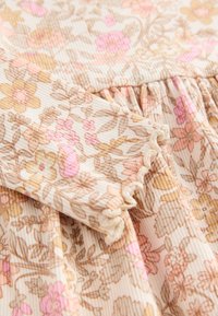 Vêtement à manches longues avec taille froncée, texture côtelée, motif floral beige, rose et marron, bordures des manches festonnées.