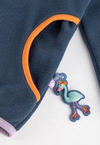 Marineblaue Fleece-Stoff mit orangefarbenen und lilafarbenen Akzenten. Verfügt über einen dekorativen Flamingo-Schlüsselanhänger mit lebendigen Details und einer Schleife am Hals.