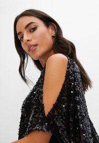Top nero con paillettes, dal design morbido, con spalle scoperte e tessuto trasparente. I paillettes riflettenti creano texture e un interesse visivo.