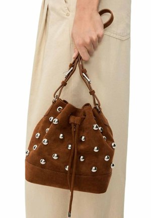 Mano che tiene una borsa a secchiello in camoscio marrone con borchie d'argento e tracolle regolabili in pelle, sfondo gonna beige.
