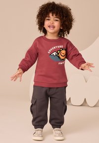 Bordo spalvos džemperis su užrašu „Nuotykiai laukia“, derinamas su pilkomis kelnėmis su kišenėmis. Modelis dėvi šviesius sportinius batelius, stovėdamas su plačiai išskėstomis rankomis.