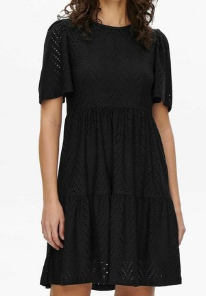 Mujer con un vestido negro de manga corta hasta la rodilla, con textura de bordado calado y falda suelta y de varias capas sobre un fondo blanco.