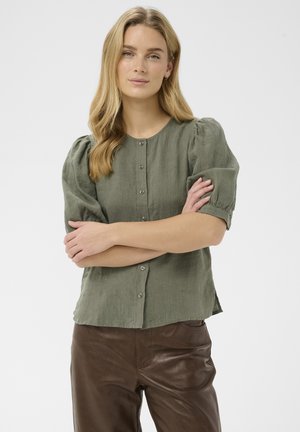 Žena s dlhými blond vlasmi, oblečená v olivovo zelenom zapínanom blúzke s nafúknutými rukávmi a tmavohnedými koženými nohavicami, so založenými rukami.