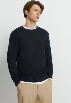 Jonge man met een marineblauwe trui over een wit overhemd en beige broek, staand met handen in de zakken tegen een effen achtergrond.