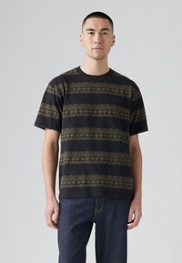 Camiseta negra de cuello redondo con patrones florales dorados horizontales. Mangas cortas y corte relajado. Llevada con jeans azul oscuro.