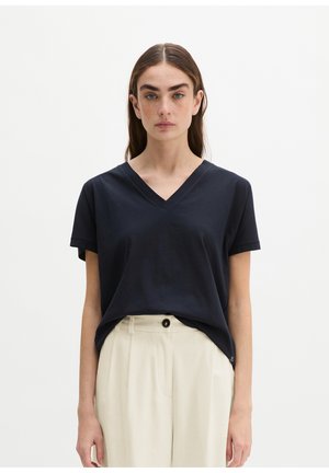 Femme aux longs cheveux bruns portant un t-shirt noir à manches courtes avec col en V, rentré dans un pantalon blanc taille haute, sur un fond uni.