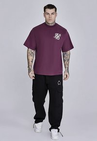 Viininpunainen lyhythihainen t-paita, jossa pyöreä pääntie ja rintakehällä kirjailtu logo, yhdistetty mustiin cargo-housuihin, joissa on sivutaskut ja logo.