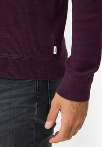 Merino wollen trui in dieppaars, met een geribbelde zoom en manchetten, en een klein wit label aan de onderzijde. Ruime pasvorm, textuur gebreid.