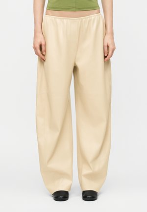 Άτομο που φορά μπεζ παντελόνια με φαρδιά μπατζάκια και ελαστική μέση, μαύρα παπούτσια, συνδυασμένα με ένα κοντό πράσινο τοπ, στέκεται σε απλό φόντο.