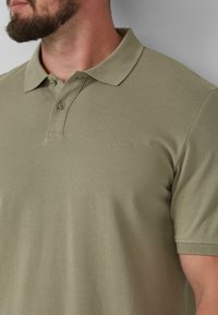 Khaki polo z teksturowanej tkaniny, z dwoma guzikami przy kołnierzyku oraz subtelnym wyszywanym logo na piersi. Krótkie rękawy.