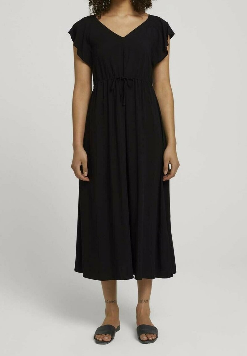Femme portant une robe midi noire à manches courtes et taille à cordon, associée à des sandales noires type slides.