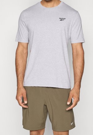 Homme portant un t-shirt Reebok gris clair et un short Reebok vert olive, debout les bras détendus le long du corps.