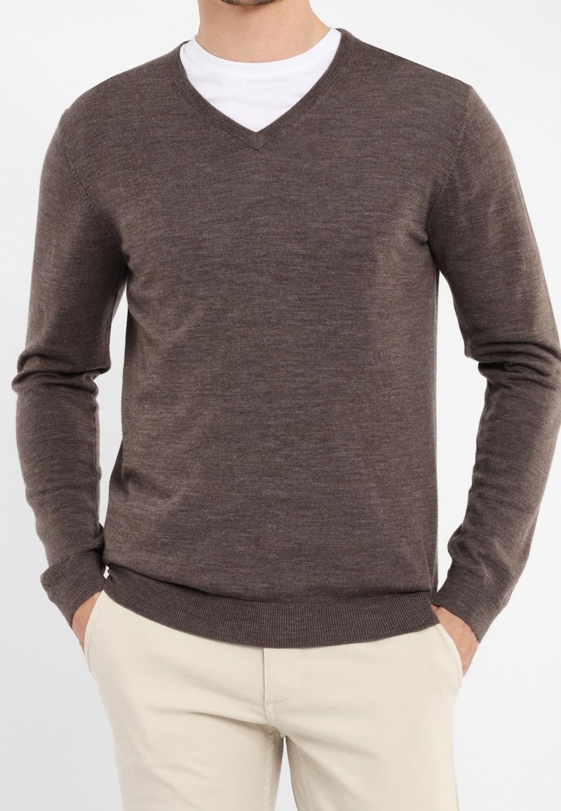 Homme portant un pull marron col en V sur une chemise blanche et un pantalon beige, les mains dans les poches, sur un fond uni.