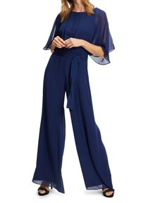 Femme portant une combinaison bleu marine à jambes larges avec des manches flottantes transparentes et une ceinture nouée à la taille, associée à des talons ouverts noirs.