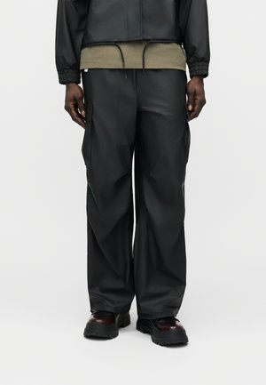 Uomo che indossa pantaloni cargo neri larghi, stivali di pelle scura e una giacca nera corta sopra una maglietta verde oliva, in piedi su uno sfondo uniforme.