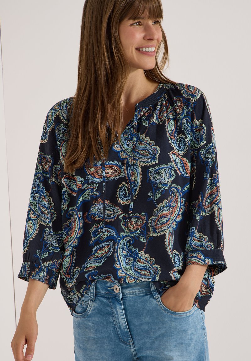 Femme souriante portant un chemisier bleu marine avec des motifs cachemire multicolores et un jean bleu clair, sa main gauche dans sa poche.