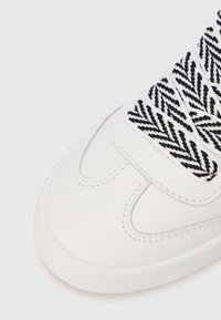 Λευκό αθλητικό παπούτσι με επάνω μέρος από λείο δέρμα, διαθέτει γλώσσα από ύφασμα με σχέδιο ψαροκόκαλο σε ασπρόμαυρο χρώμα και αντίθετες ραφές ως λεπτομέρεια.