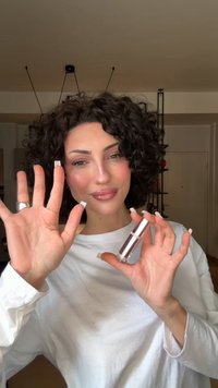 Clear glass lip gloss container with a silver cap held in raised hand. Subject wears a white long-sleeve shirt and has curly hair.