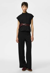 Mladić stoji noseći crni top bez rukava, široke crne hlače, široki smeđi remen s torbicom i grimizne tenisice.